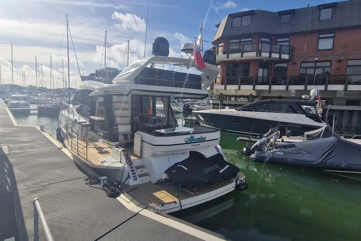 A luxury yacht named "Mannie" docked at a marina, surrounded by other boats and a waterfront building in bright sunlight.